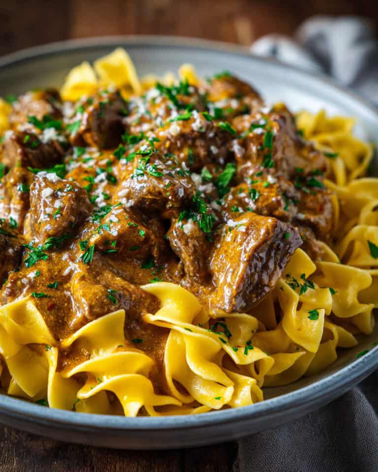 Rich beef stew served over homemade egg noodles, topped with parsley, on a gray plate.